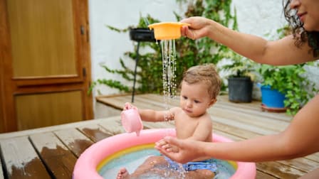 Baby playing in a small inflatable pool while an adult pours water from a toy above.