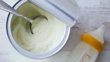 overhead view of open container of baby formula with measuring spoon, baby bottle blurred in background