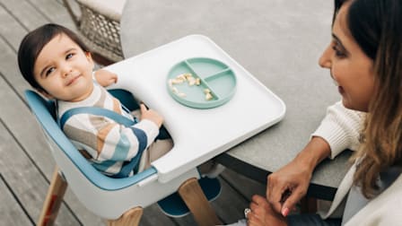baby in striped sweater sitting in Uppababy Ciro high chair at round table, with adult smiling at them in corner of frame