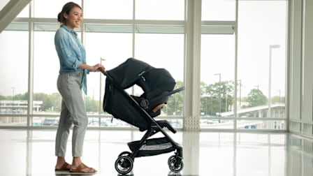 person wearing denim shirt and pants pushing Nuna Triv Next + Pipa Aire RX Travel System through airport with large glass windows behind them