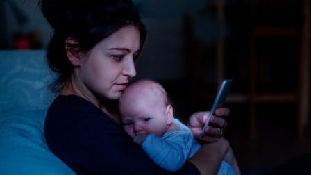 Mother holding an awake infant in the middle of the night while looking at her phone.