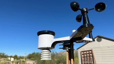 Ambient Weather weather station on wooden post with blue sky, desert landscape, and building in background