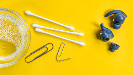 blue earbuds with three cotton swaps, two paperclips, and small bowl of soapy water on yellow background