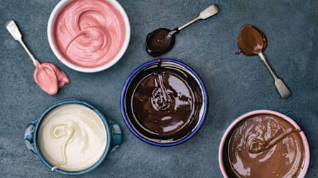overhead view of four different types of melted chocolate in bowls (white, ruby, dark, and milk) with spoons covered in chocolate next to bowls