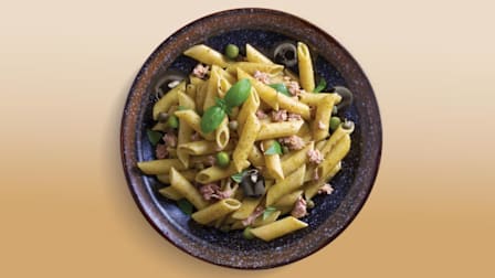 overhead view of bowl of penne with basil, peas, tuna fish, and black olives on brown background