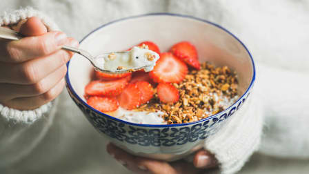 close up of person in white sweater holding blue and white ceramic bowl with sliced strawberries, granola, and yogurt