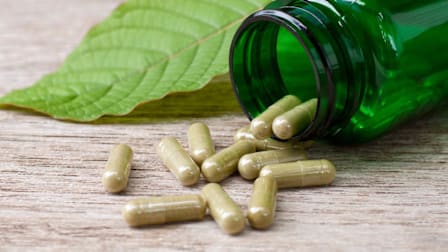 Kratom supplement capsules spilling onto a surface from a pill bottle next to a kratom leaf.