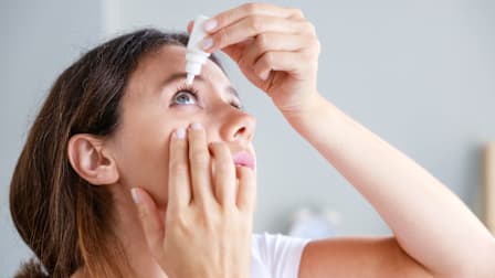 close up of person using eye drops, one hand on cheek and the other holding eye dropper container above eye