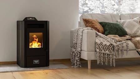 United States Stove Modern Wood Pellet Stove with 50lb Hopper in living room with beige couch with blankets and pillows, snow on trees through window behind couch