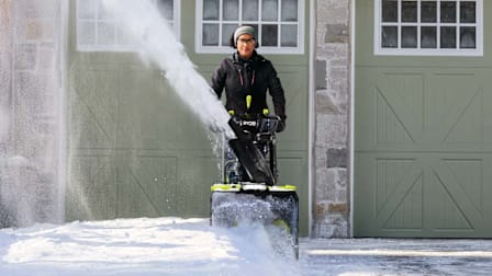 A woman using a Ryobi RY408150 Two-Stage Battery Snow Blower to clear snow off a residential driveway.