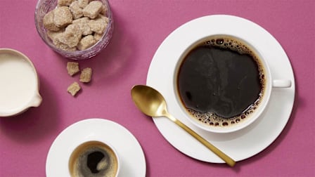 A top-down view of a cup of coffee, an espresso, a creamer pitcher, and a jar of brown sugar cubes on a purple surface.