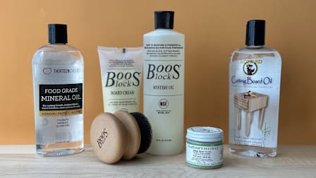Thirteen Chefs Food Grade Mineral Oil, Boos Block Board Cream and Mystery Oil with Brush set, Clapham's Beeswax, and Howard Cutting Board Oil on wooden cutting board with light brown background