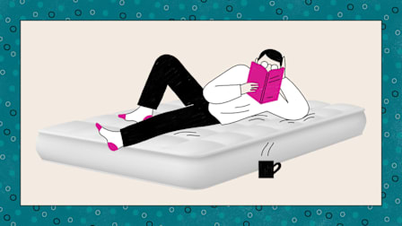 A person relaxing on a mattress, reading a book with a steaming mug nearby.