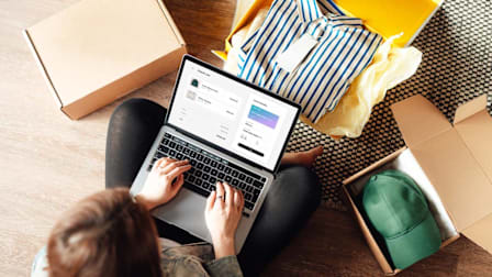 A person sits on the floor using a laptop to shop online, surrounded by open boxes with clothing items including a striped shirt and a green cap.