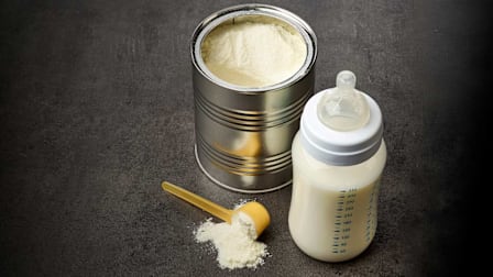 Still life of a can of baby formula with a baby bottle and scoop of formula.