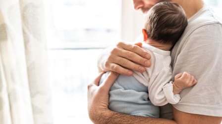 Man holding an infant to his chest next to a window.
