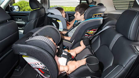 A side view shows three children secured in car seats arranged side-by-side across a vehicle'a back bench seat. In the foreground, an infant lies in a rear-facing car seat, while a young boy sits in a forward-facing car seat in the middle. On the far side, a young girl is secured in a rear-facing car seat.