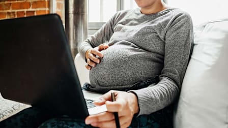 A pregnant person in a grey sweater sits on a white couch while using a laptop computer. One hand rests gently on their baby bump while the other holds a pen near a laptop thats placed on their lap.