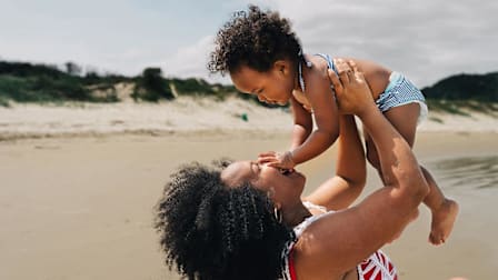 A smiling mother playfully lifts her toddler into the air on a beach with sand dunes in the background.