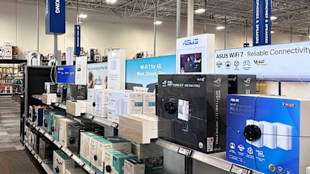 Interior of an electronics store showing shelves stocked with various router brands with a vertical sign saying "WI-FI & Networking".
