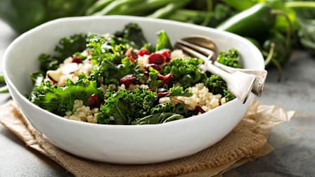 white ceramic salad bowl with kale, quinoa, and dried cranberries sitting on burlap with greens and peppers out of focus in background