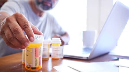 person reaching for prescription bottle with other bottles nearby, sitting in front of laptop