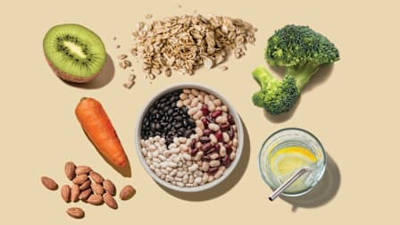 half a kiwi, carrot, almonds, glass of water with metal straw, broccoli floret, oatmeal pile, and bowl of beans on beige background