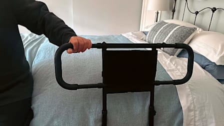 A person's hand grips a black, U-shaped metal bed assist rail that is positioned against the side of a bed in a residential bedroom.