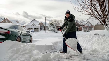 person shoveling deep snow from driveway in suburban neighborhood with other houses in background