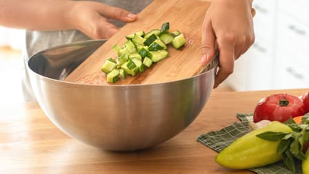 Person sliding cucumbers from wooden cutting board to metal bowl in kitchen