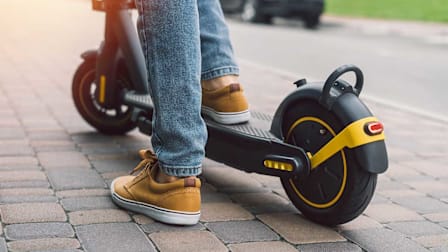 A close-up shot of a person's lower legs and feet while riding a black electric scooter on a paved cobblestone sidewalk.