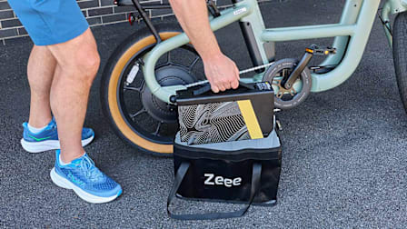 A person is shown placing a large, patterned e-bike battery into a black "Zeee" brand bag on the ground next to a mint green electric bicycle.