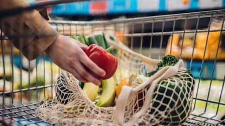 Person placing a red pepper in their shopping cart along with bananas, an orange and a head of broccoli.