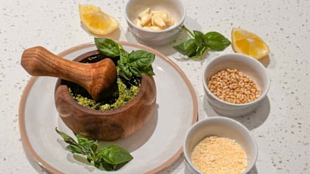Homemade pesto in a wooden mortar and pestle, surrounded by fresh basil, garlic, pine nuts, Parmesan cheese, and lemon wedges.