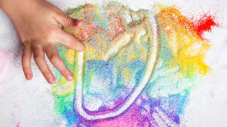 Child's hand creating a rainbow shape in a mixture of colorful sand.