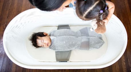 A baby sleeping in a Happiest Baby Snoo bassinet with his mother and sister looking at him