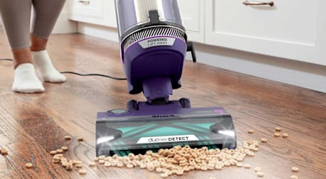 A person using the Shark PowerDetect AZ4002 upright vacuum to clean up spilled dry cereal from a kitchen floor.