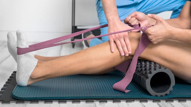 Person using a resistance belt and foam roller on fitness mat while physical therapist offers guidance