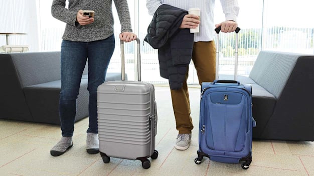 low view of two people with carry-on luggage, holding coffee container and phone, with couches and windows behind them