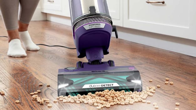 A person using the Shark PowerDetect AZ4002 upright vacuum to clean up spilled dry cereal from a kitchen floor.