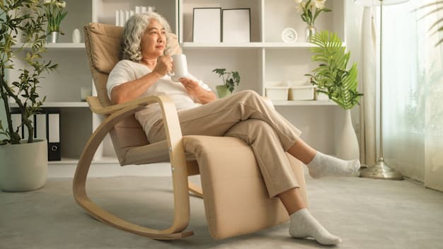 Person sitting in living room in cushioned chair holding mug, with shelves and plants in background