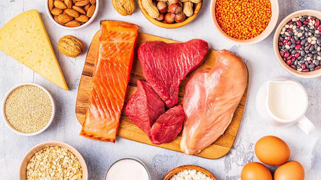overhead view of different types of protein: raw salmon, beef, and chicken on wooden cutting board, nuts, various whole grains and beans in bowls, cheese, eggs, yogurt, and milk on light grey background