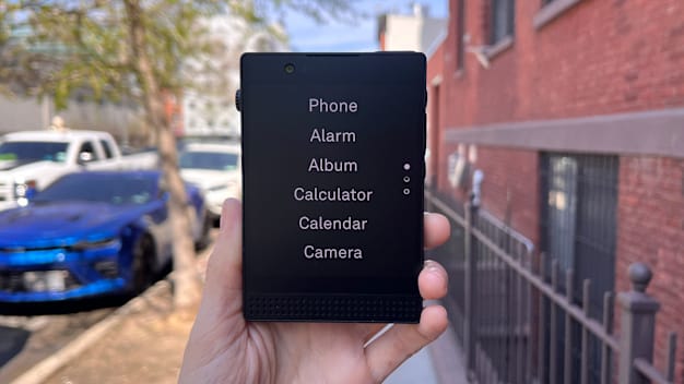 handing hold Light Phone III with navigation menu on the screen, cars and brick building in the background