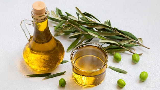 glass olive oil pitcher with bowl of olive oil, olives, and olive greens