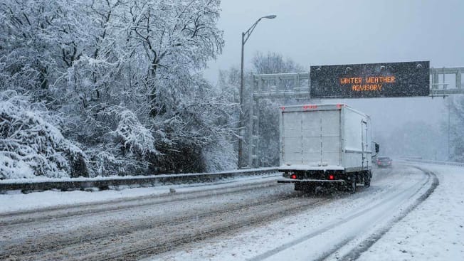 A truck driving on a highway during a winter storm with a illuminated highway sign overhead advising of a Winer Weather Advisory.
