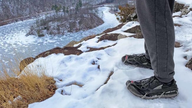 detail of person's legs and feet wearing sneakers and ice cleats, standing at edge of hill with river in background
