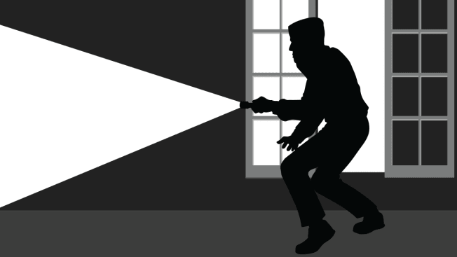 An illustration of a burglar using a flashlight after having broken into a home through a window.