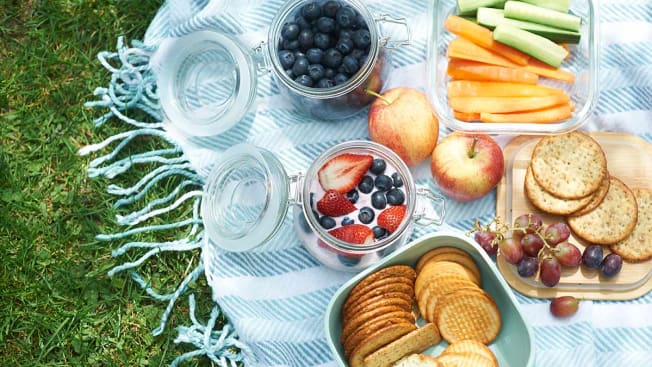 Closeup of a picnic spread with fruit, veggies and crackers on a blanket in the grass.