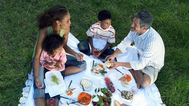 overhead view of parents and two children sitting on white blanket with picnic foods, surrounded by grass