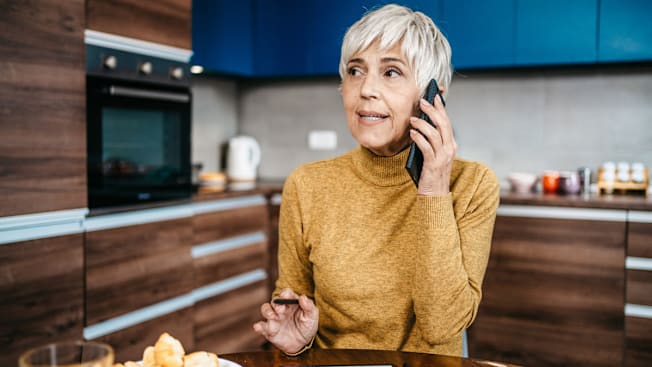 person in yellow turtleneck talking on phone while sitting at table, kitchen cabinets and wall oven blurred in background